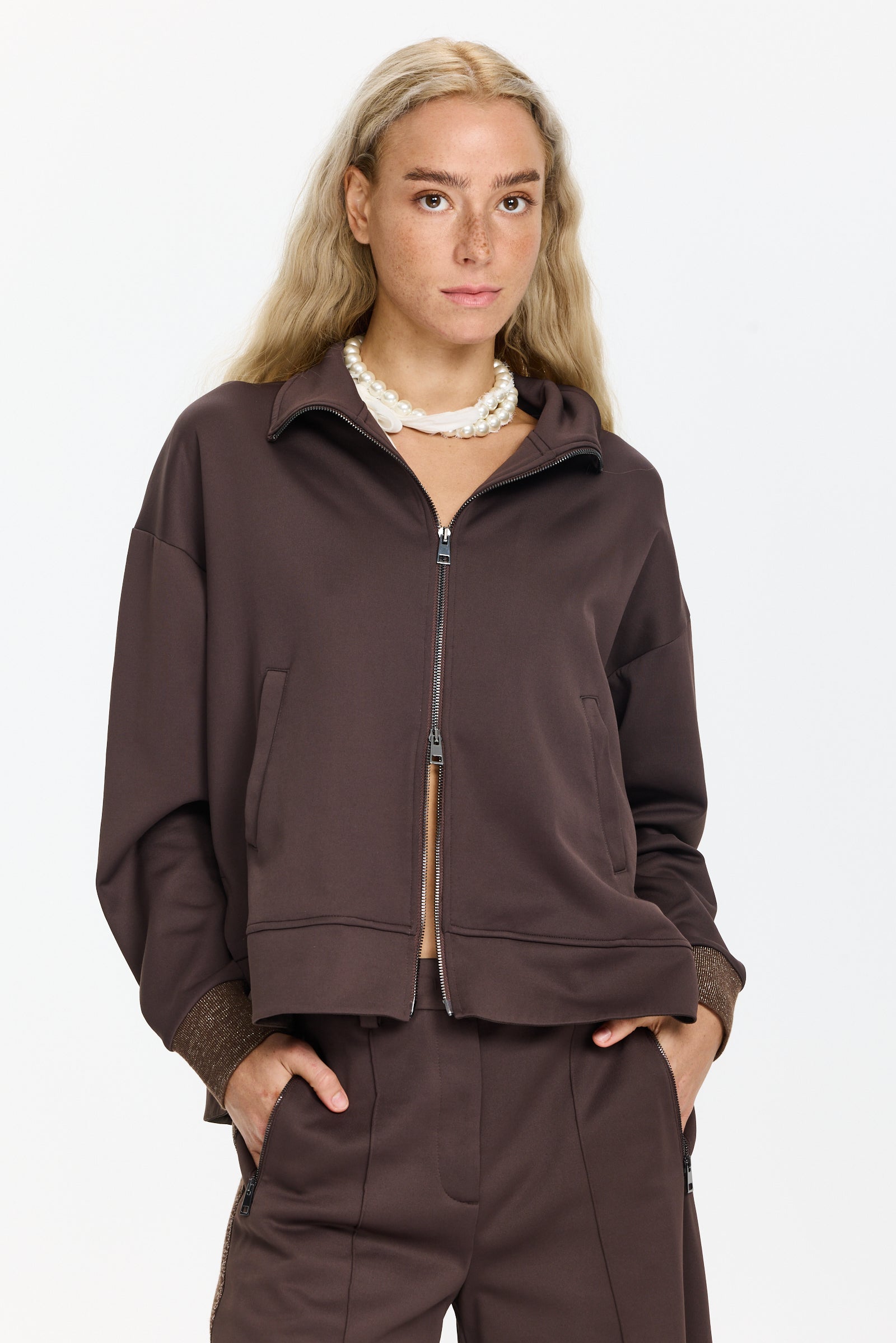סווטשירט בצבע חום כהה עם צווארון גבוה, רוכסן כפול, כיסים קדמיים וכוכב בגב, קצוות השרוולים נוצציםהדוגמנית בגובה 1.75 ולובשת מידה 40 של Lorena Antoniazzi